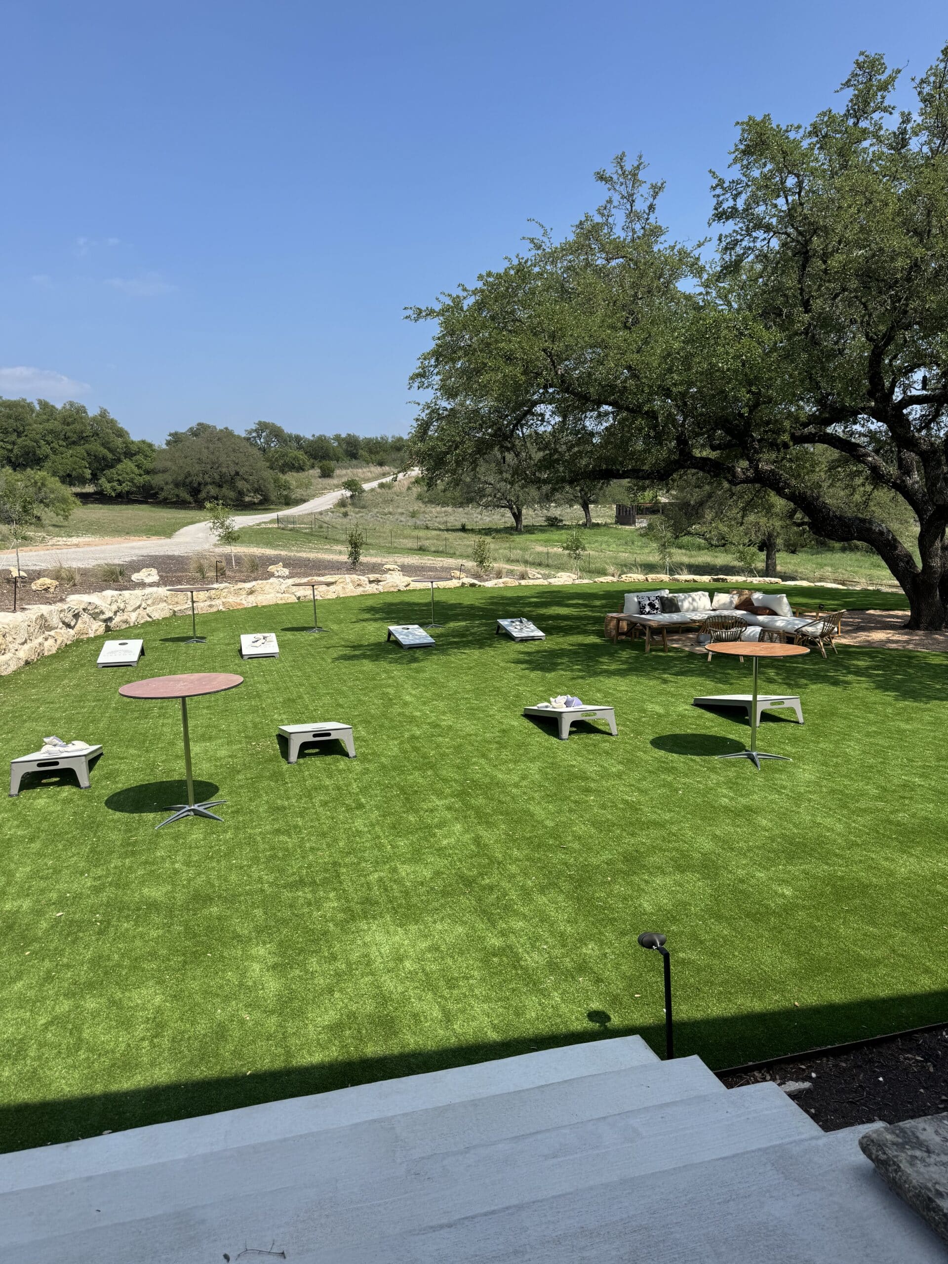 Camp Hideaway Spicewood lawn with cornhole, cocktail tables, and live oak canopy — outdoor group activity space with Hill Country terrain in the background