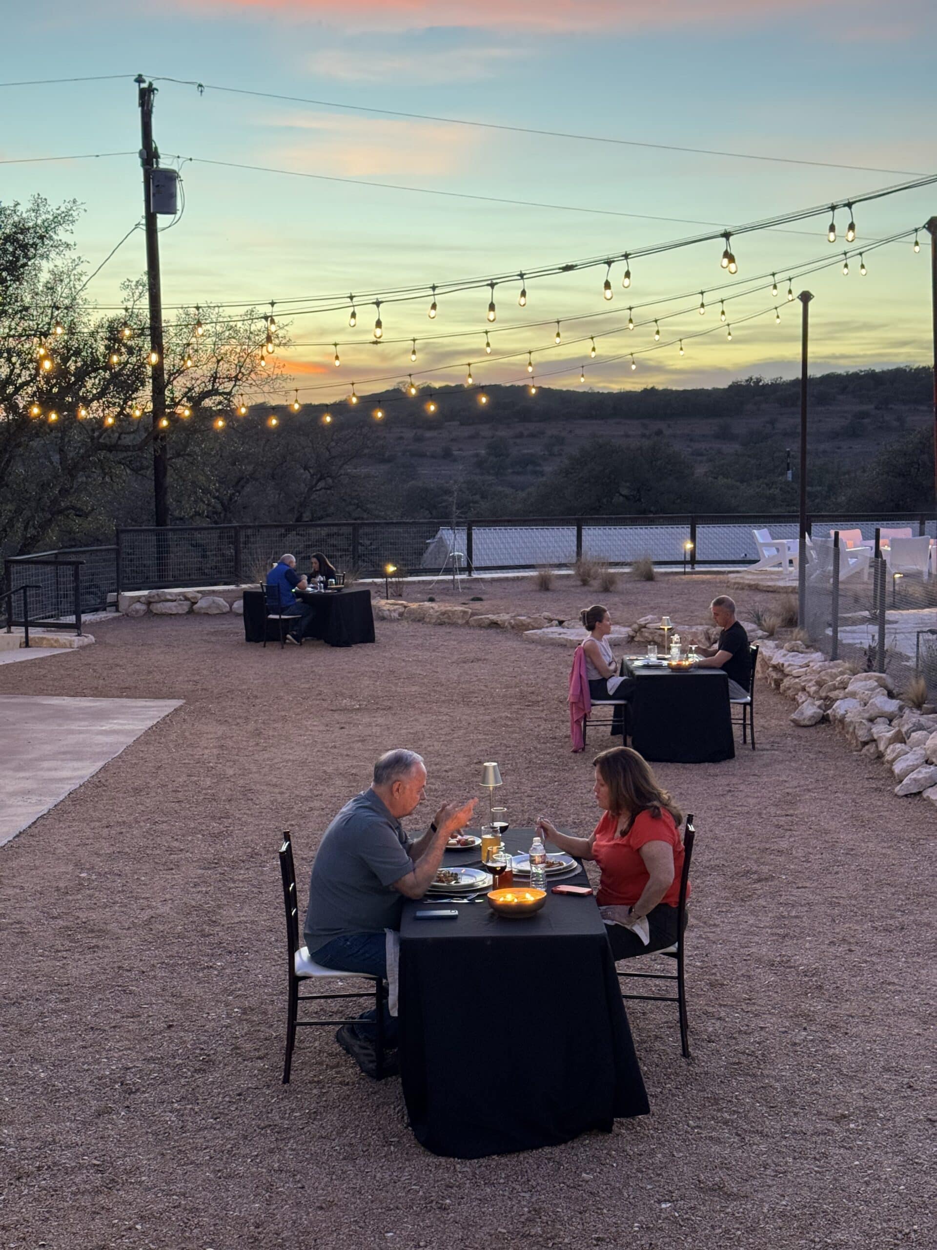 Outdoor courtyard dining at dusk at Camp Hideaway Spicewood — string lights, Hill Country terrain, groups gathered at tables under the Texas sunset